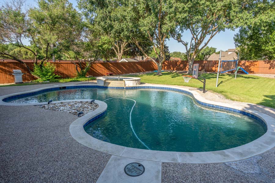 Weekly Pool Cleaning Service by Flower Mound Pool Care and Maintenance. Image of a jacuzzi in the background taken from an angle in back of house. Image shows basketball hoop and a volley ball net in the back.