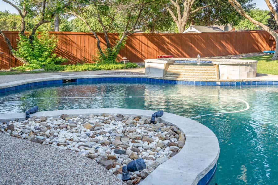 Weekly Pool Cleaning Service by Flower Mound Pool Care and Maintenance. Image of a jacuzzi in the background taken from a front angle in back of house. Image show decorative rock and lighting.