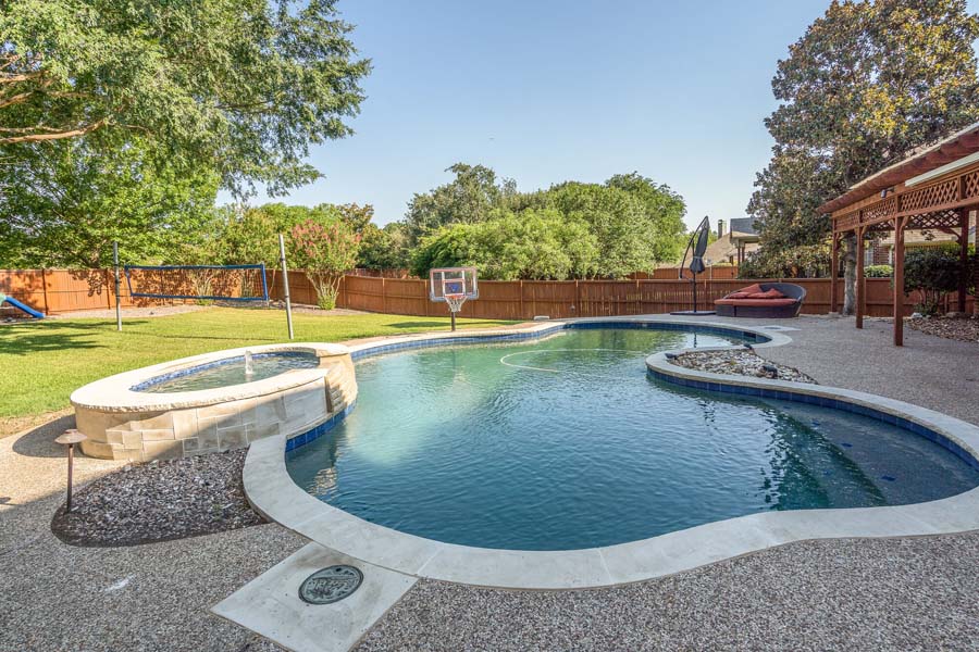 Weekly Pool Cleaning Service by Flower Mound Pool Care and Maintenance. Image of a nicely remodeled pool and jacuzzi in the foreground taken from a side angle in back of house.