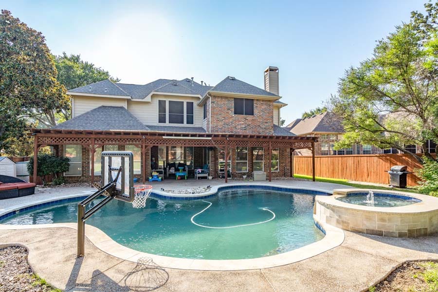Weekly Pool Cleaning Service from Flower Mound Pool Care. Image taken from back of house, with sun shade, and table and chairs. House has brick facade. More lounging chairs and stools in front of windows