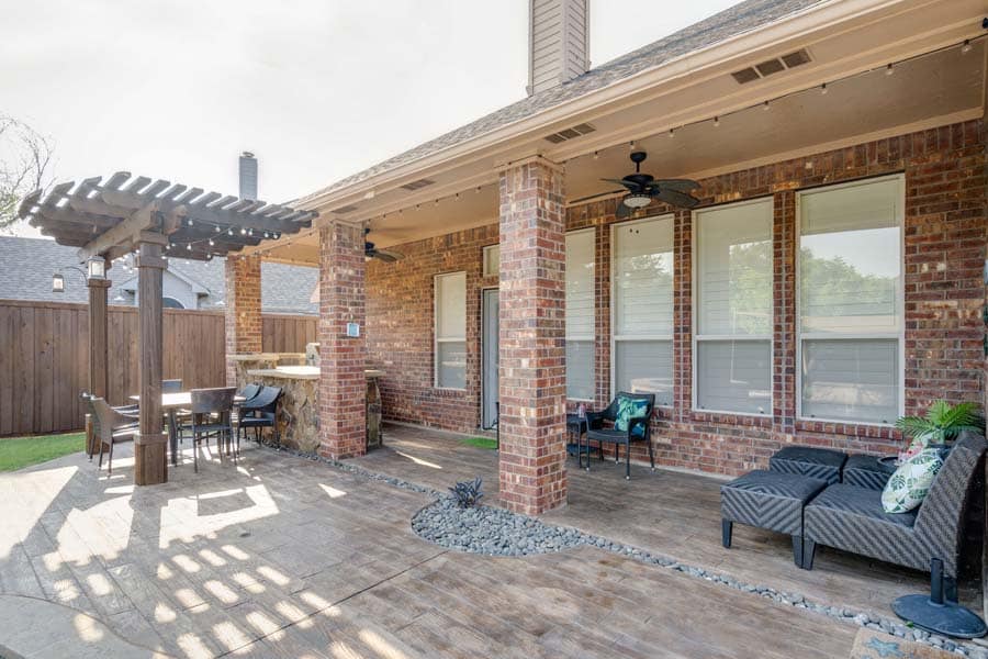 Weekly Pool Cleaning Service from Flower Mound Pool Care. Image taken from an angle of patio, with sun shade, and table and chairs. House has brick facade. More lounging chairs and stools in front of windows