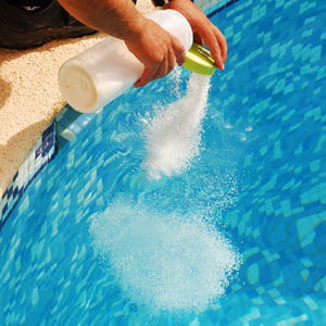 Photo showing pool technician adding chemicals to a pool. Water is blue.
