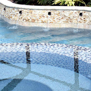 Picture of the edge of a pool with water feature where the water is pouring into the pool thourgh tubes. Water is blue, side of pool is white, tan and brown small tiles.
