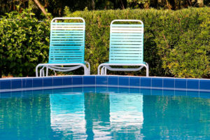 Picture of a pool with blue tire and two light blue lounge chairs are shown in the center. The pool is surrounded by a shrubbery all along the back side.
