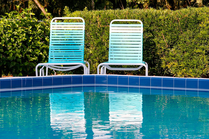 Picture of a pool with blue tire and two light blue lounge chairs are shown in the center. The pool is surrounded by a shrubbery all along the back side.